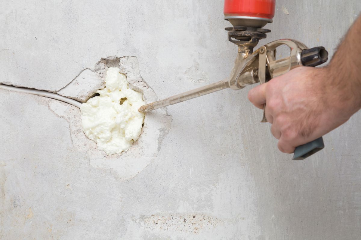 Young man hand using spray gun and filling hole with construction foam in concrete wall where was old outlet. Closeup. Repair work of home. Renovation process. Side view.