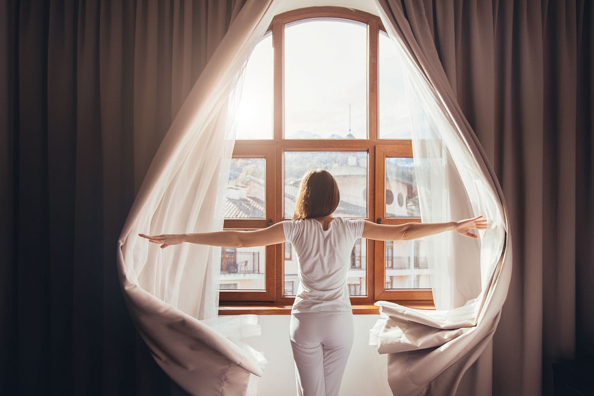 young fit woman in home clothes opens curtains of bedroom window