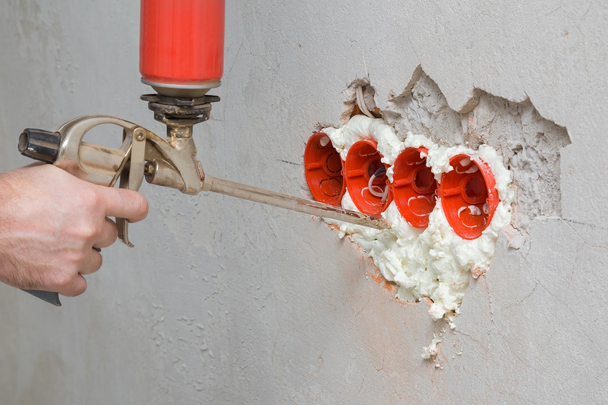 Hand using spray gun and filling gap with construction foam between new plastic red electric outlet and concrete wall