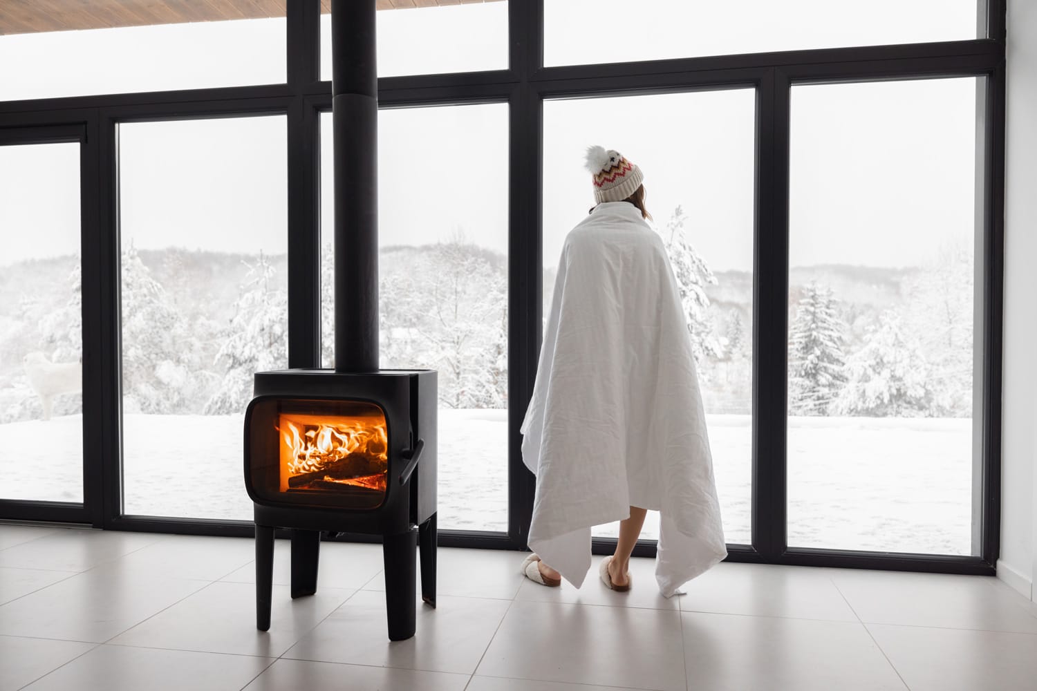 Woman enjoys winter time at home with burning fireplace and panoramic window, looking outside on snowy landscape. Concept of winter mood and comfort at home. Idea of winter vacation in the mountains