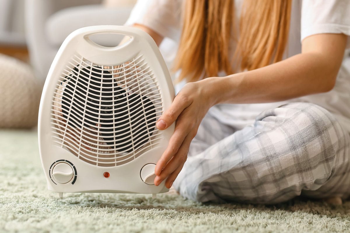 Woman with electric fan heater sitting on carpet at home