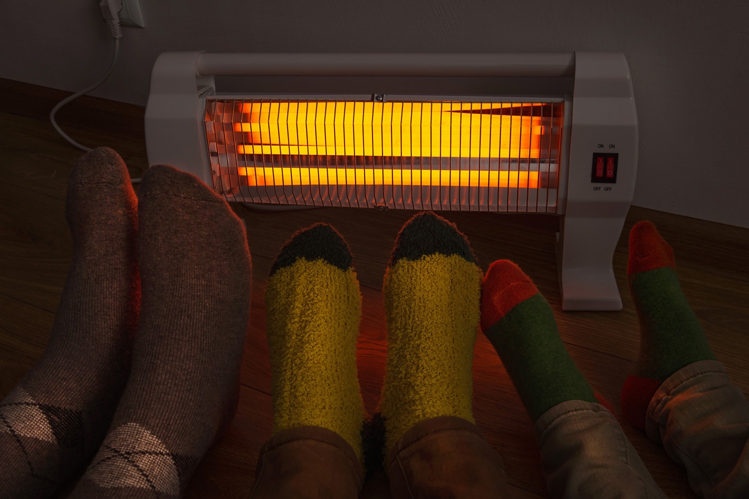 Young family together warms cold legs near an infrared electric heater in darkness at home.