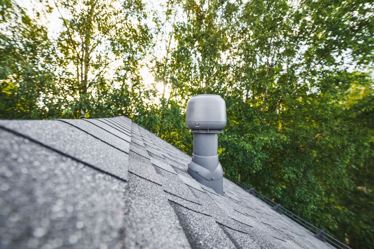 renovated roof ridge with shingles roof-tiles of a house and ventilation pipe, green trees background with sunlight