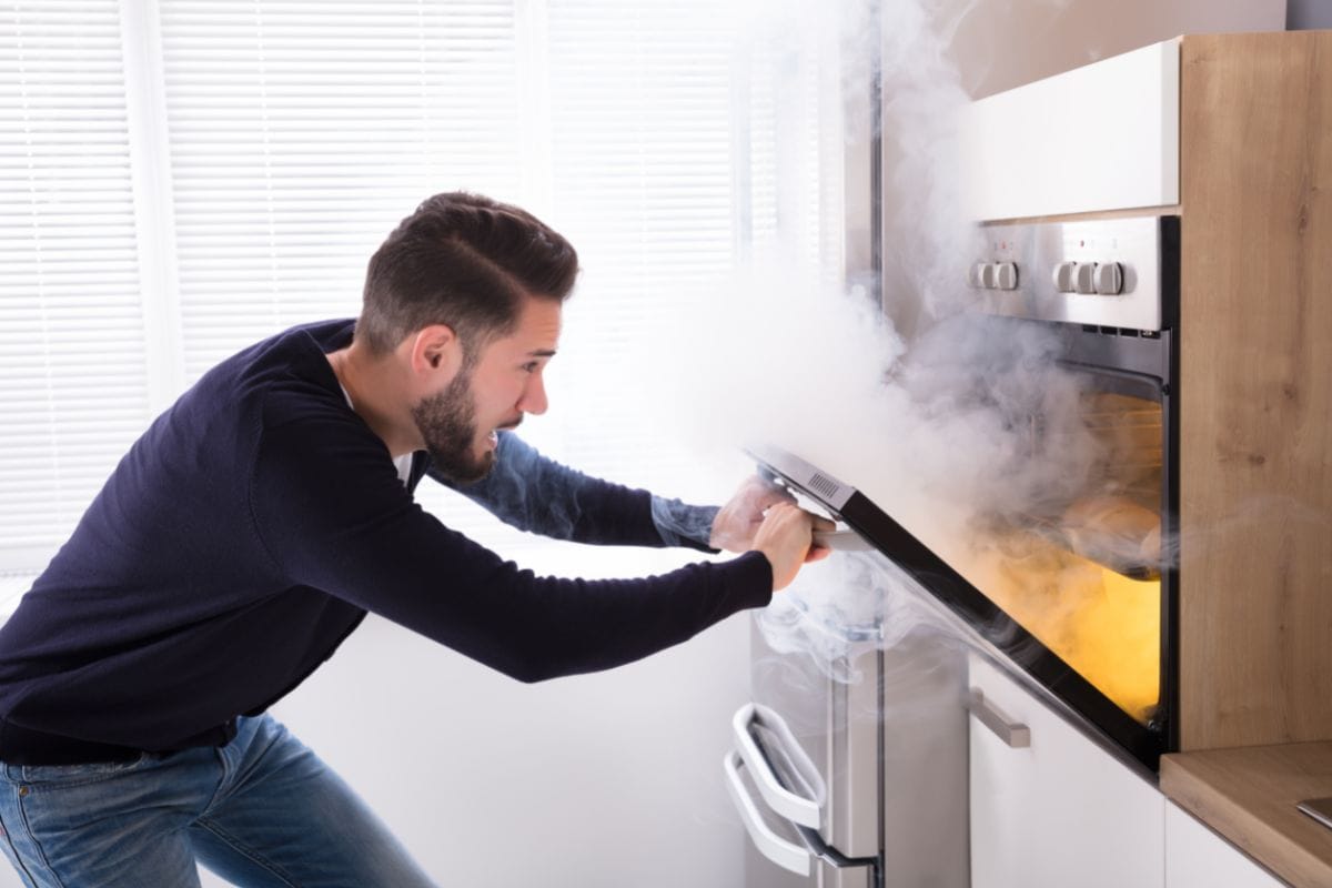 Shocked Man Looking At Burnt Cookies With Smoke Coming From Oven