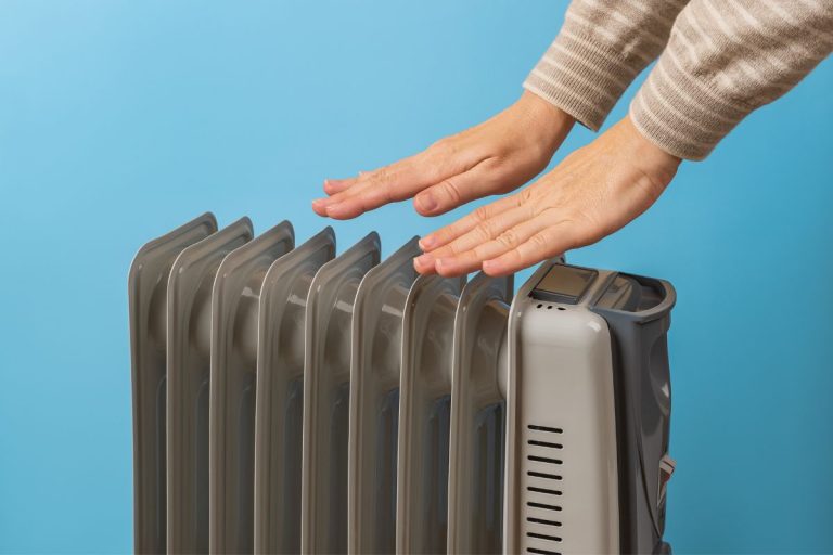 Woman warms her hands near hot oil filled electric heater against blue background. Using 9 fins space heater. Portable household appliance for heating home
