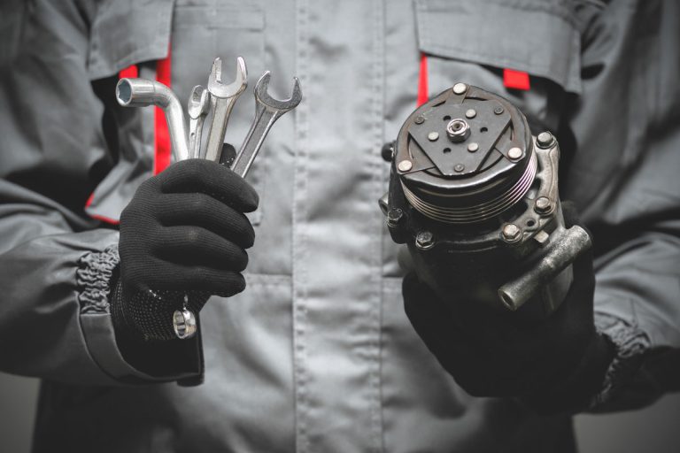 mechanic holding the tools in the right hand and in the left hand is the ac compressor, Where Is The AC Compressor Located In A Car? [Detailed Guide]