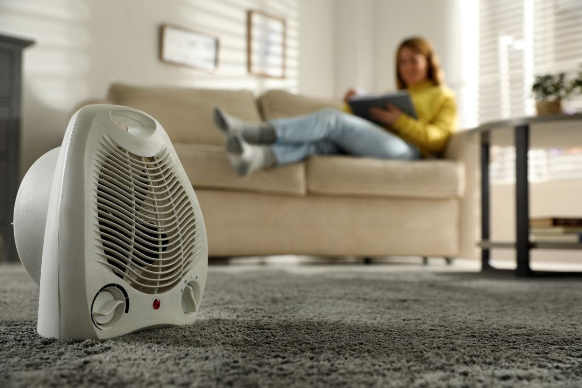 Woman reading book in living room, focus on electric fan heater.